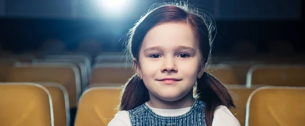 Panoramic shot of cute brunette child sitting in cinema and looking at camera — Stock Photo