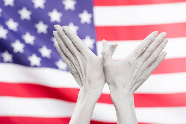 Vue recadrée des mains féminines peintes en blanc montrant un signe en forme d'oiseau près du drapeau américain — Photo de stock