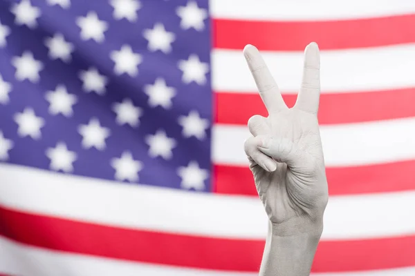 Vista recortada de la mano femenina pintada de blanco que muestra signo de paz cerca de la bandera americana - foto de stock