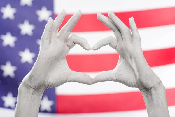 Cropped view of female hands painted in white showing heard-shaped sign near flag of america — Stock Photo