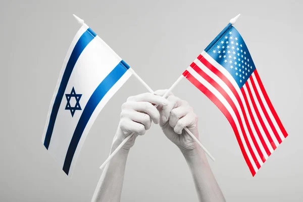 Vista recortada de manos femeninas pintadas de blanco sosteniendo banderas cruzadas americanas e israelíes aisladas en gris - foto de stock