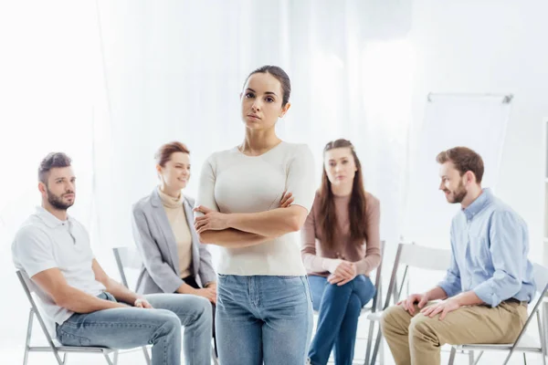 Frau mit verschränkten Armen blickt in die Kamera, während die Menschen während der Gruppentherapie auf Stühlen sitzen — Stockfoto