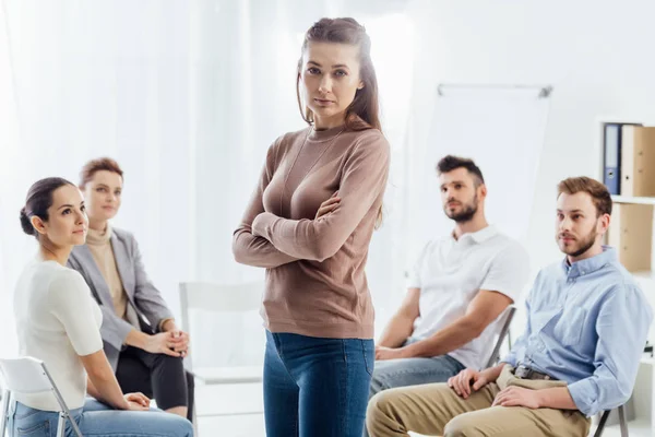 Frau mit verschränkten Armen blickt in die Kamera, während die Menschen während der Gruppentherapie auf Stühlen sitzen — Stockfoto