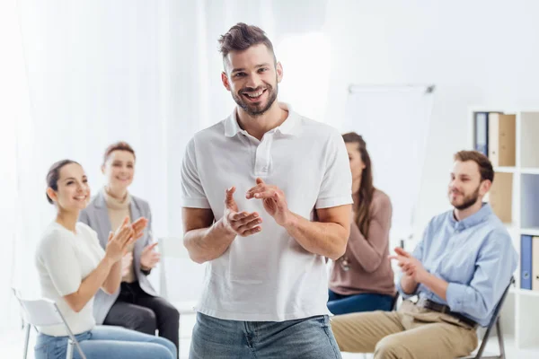 Hombre mirando a la cámara y aplaudiendo mientras la gente se sienta durante la sesión de terapia de grupo - foto de stock