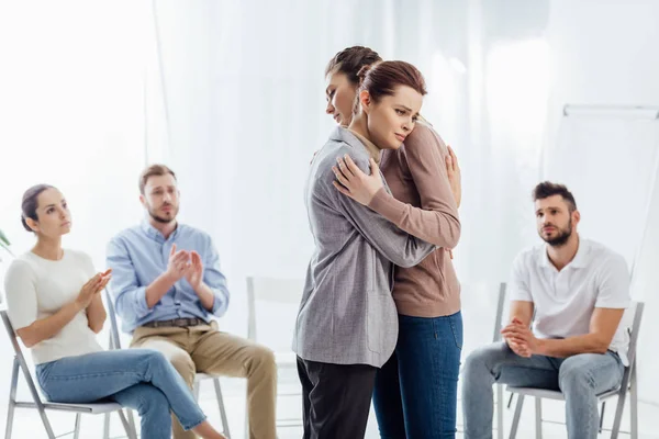 Frauen umarmen sich, während eine Gruppe von Menschen sitzt und während der Therapiesitzung applaudiert — Stockfoto