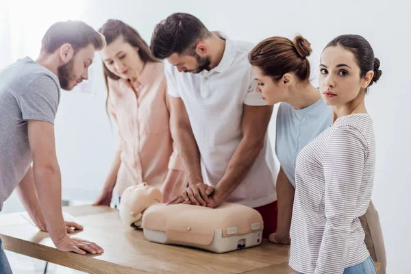 Mujer mirando a la cámara mientras grupo de personas realizando cpr en maniquí durante el entrenamiento de primeros auxilios - foto de stock