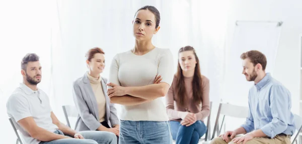 Foto panorámica de la mujer con los brazos cruzados mirando a la cámara mientras las personas sentadas durante la sesión de terapia de grupo - foto de stock