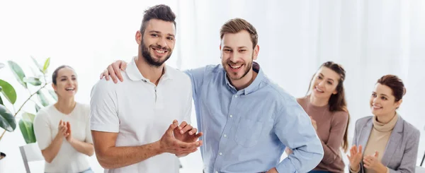 Two smiling men looking at camera while people sitting and applauding during group therapy session — Stock Photo