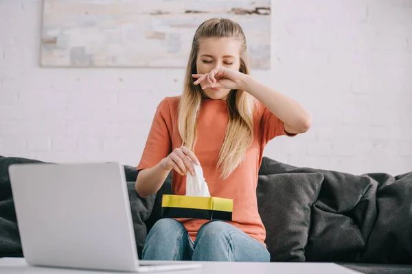 Blonde woman sneezing while holding tissue box near laptop — Stock Photo