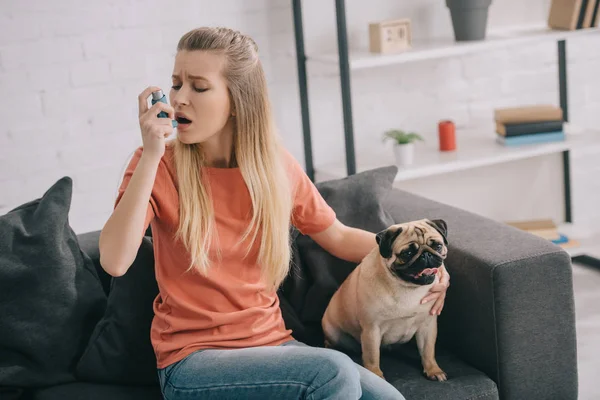 Attraktive blonde Frau allergisch gegen Hund mit Inhalator, während sie in der Nähe von Mops zu Hause sitzt — Stockfoto