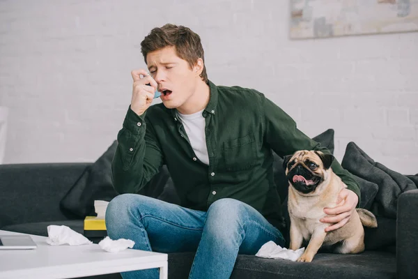 Hombre guapo alérgico al perro usando inhalador mientras está sentado en el sofá con pug - foto de stock