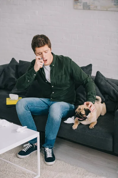 Hombre guapo alérgico al perro usando inhalador mientras está sentado en el sofá y mirando pug - foto de stock