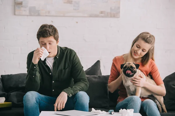 Attrayant blonde femme avec carlin près éternuer homme allergique au chien sur canapé — Photo de stock