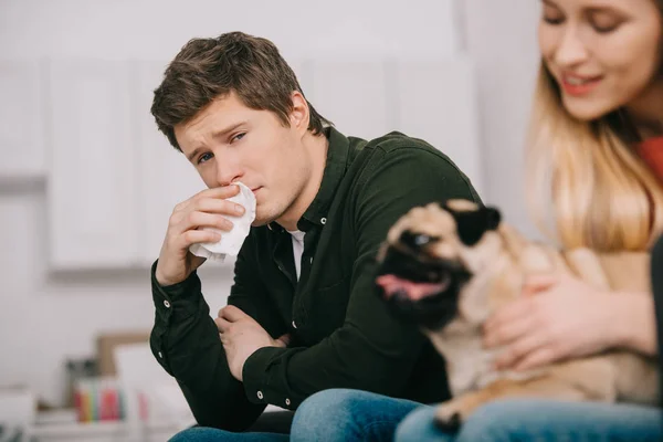 Selective focus of man allergic to dog holding tissue and sitting with cheerful woman with pug — Stock Photo