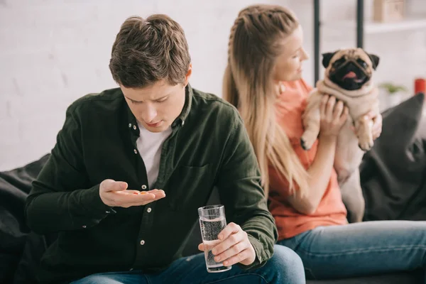 Selective focus of handsome man allergic to dog holding glass of water and looking at pills near blonde woman with pug — Stock Photo