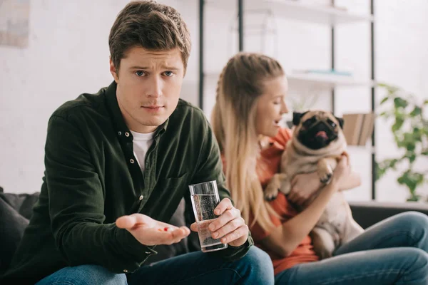 Selektiver Fokus eines gutaussehenden Mannes, der allergisch auf einen Hund reagiert, der ein Glas Wasser und Pillen in der Nähe einer blonden Frau mit Mops hält — Stockfoto