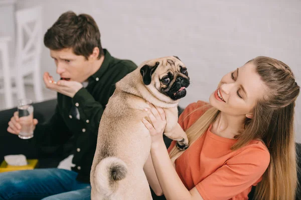 Enfoque selectivo de la mujer feliz con pug cerca de hombre guapo alérgico al perro sosteniendo un vaso de agua y tomando pastillas - foto de stock