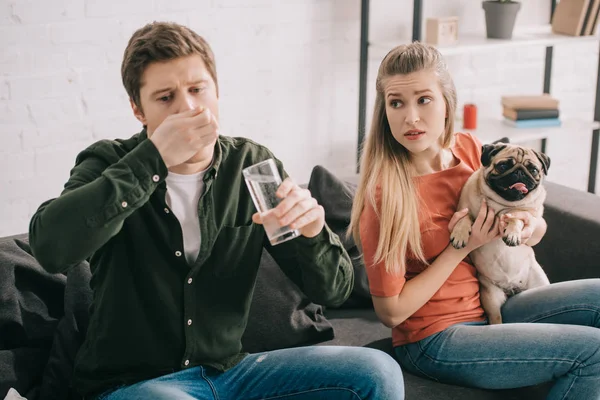 Selective focus of blonde woman with pug looking at handsome man allergic to dog holding glass of water and taking pills — Stock Photo