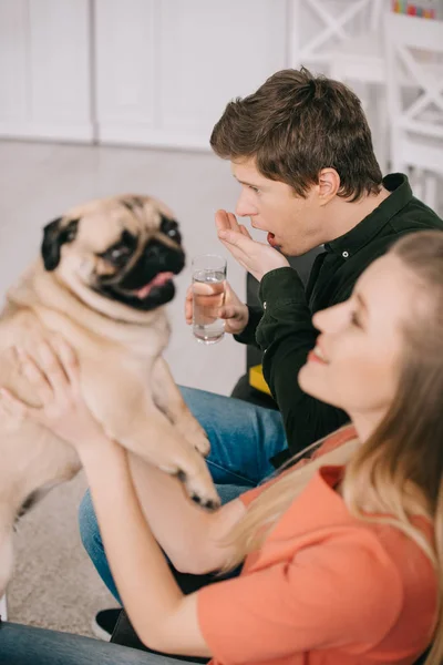 Селективное внимание красивого мужчины с аллергией на собаку, держащую стакан воды и принимающую таблетки рядом с блондинкой с мопсом — стоковое фото
