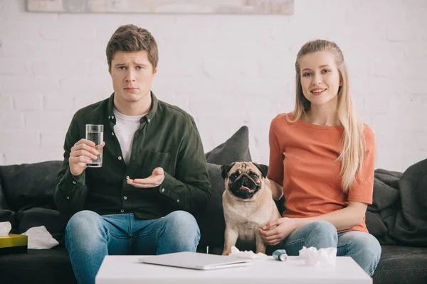 Красивий чоловік алергія на собаку тримає склянку води і таблетки біля веселої жінки з милим мопсом — стокове фото