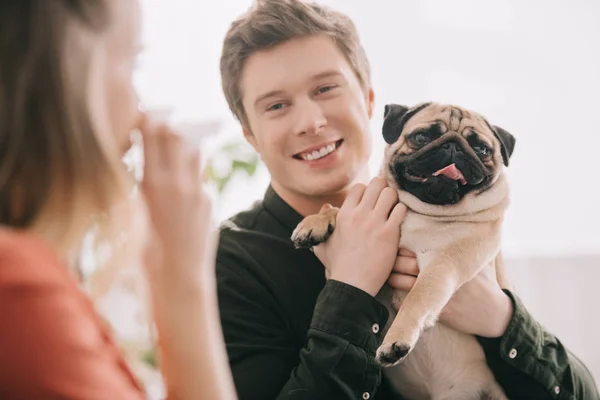 Selective focus of cheerful handsome man holding pug and looking at woman allergic to dog with tissue — Stock Photo