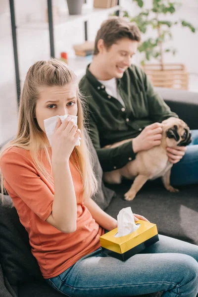 Selective focus of sneezing woman allergic to dog looking at camera near man with pug — Stock Photo