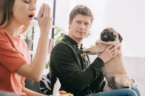 Selektiver Fokus eines gutaussehenden Mannes, der einen süßen Mops in der Hand hält und ein Mädchen ansieht, das mit Inhalator allergisch auf Hunde reagiert — Stockfoto