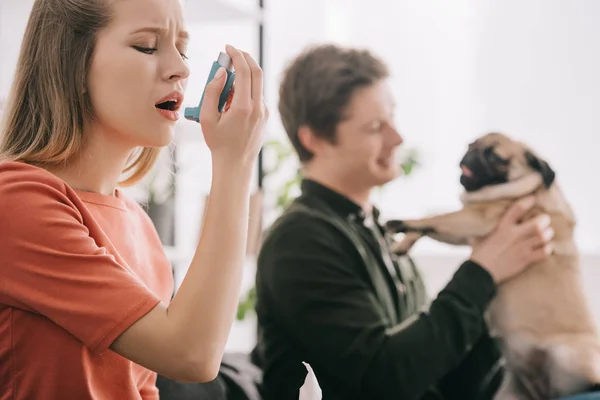 Foyer sélectif de la femme blonde attrayante allergique au chien en utilisant inhalateur près de l'homme avec carlin — Photo de stock