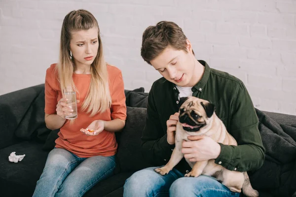 Attractive blonde woman allergic to dog holding glass of water and pills near man with pug — Stock Photo