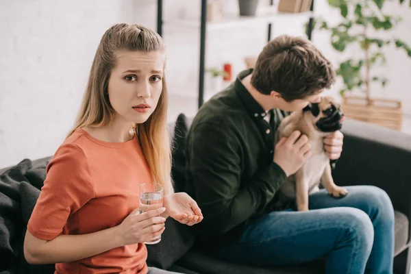 Selektiver Fokus einer verärgerten Frau, die allergisch auf Hundehaltungen mit Pillen und einem Glas Wasser in der Nähe eines Mannes mit Mops reagiert — Stockfoto