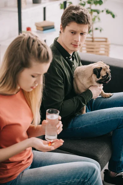 Foco seletivo do homem com pug e olhando para a mulher alérgica ao cão segurando vidro de água e pílulas — Fotografia de Stock