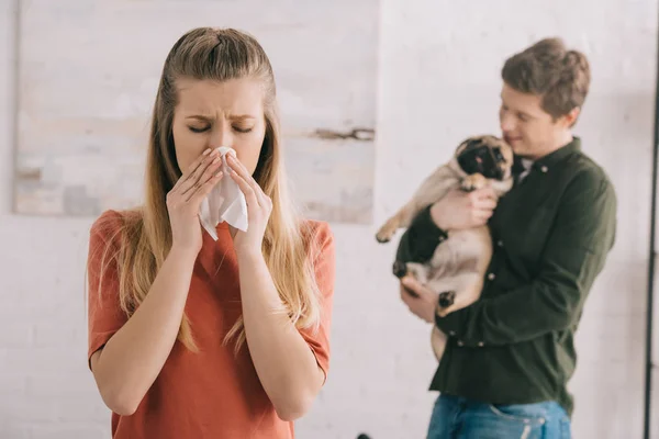 Selective focus of blonde girl allergic to dog sneezing in white tissue near man with cute pet — Stock Photo