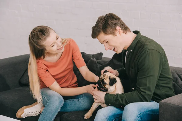 Cheerful blonde girl and happy man sitting with cute pug dog on sofa — Stock Photo