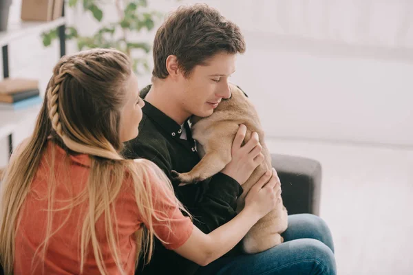 Hombre feliz sosteniendo en brazos perro lindo pug mientras está sentado en el sofá cerca de chica rubia - foto de stock