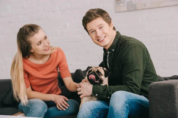 Fröhliche blonde Frau schaut glücklichen Mann an, während sie mit süßem Mops auf Sofa sitzt — Stockfoto