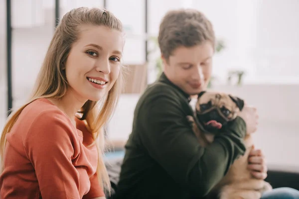 Selektiver Fokus der fröhlichen blonden Frau, die in die Kamera schaut, in der Nähe eines glücklichen Mannes mit niedlichem Mops-Hund — Stockfoto