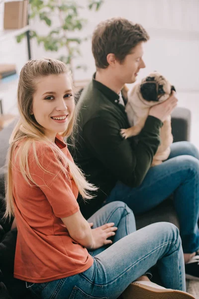 Вибірковий фокус щасливої блондинки, дивлячись на камеру біля щасливого чоловіка з милим мопсом собакою — стокове фото