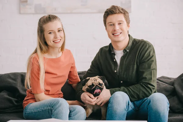 Feliz rubia mujer y guapo alegre hombre sentado con lindo perro pug en sofá - foto de stock