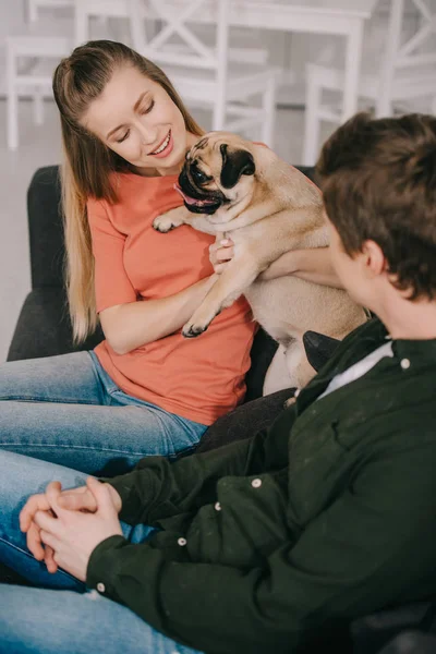 Selective focus of happy blonde woman holding in arms cute pug dog near man sitting on sofa — Stock Photo