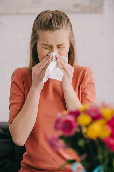 Fuoco selettivo della donna bionda con allergia al polline starnutisce nel tessuto con gli occhi chiusi vicino ai fiori — Foto stock