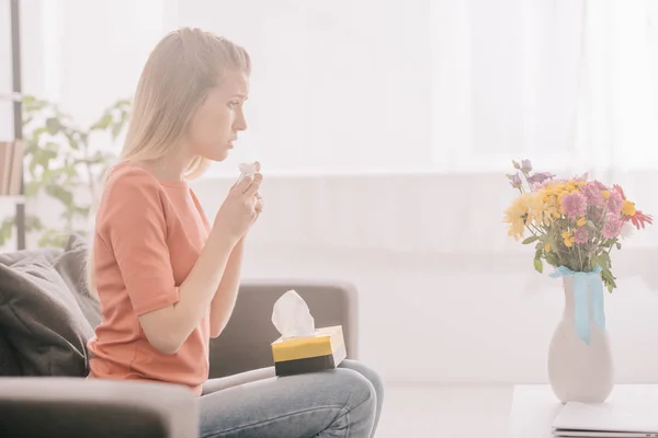 Vue latérale de femme blonde triste avec allergie au pollen tenant des tissus et regardant des fleurs dans un vase — Photo de stock