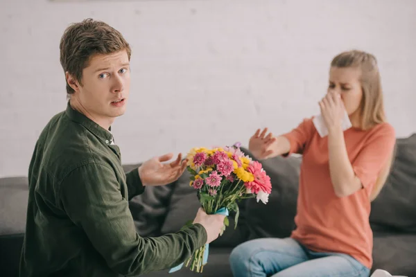 Upset man holding flowers and looking at camera near blonde woman with pollen allergy sneezing in tissue — Stock Photo