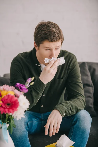 Bel homme avec des éternuements d'allergie au pollen près des fleurs tout en étant assis sur le canapé — Photo de stock