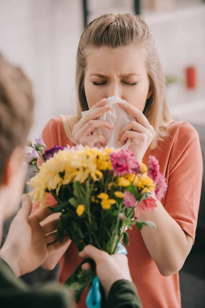 Селективный фокус чихающей блондинки с аллергией на пыльцу рядом с мужчиной с цветами — стоковое фото