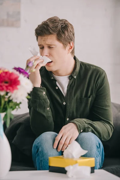 Bel homme avec des éternuements d'allergie au pollen dans les tissus près des fleurs tout en étant assis sur le canapé — Photo de stock