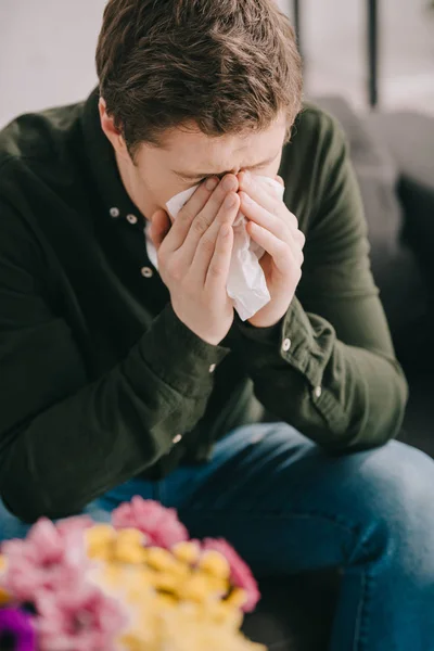 Man with pollen allergy covering face while sneezing in tissue near flowers — Stock Photo