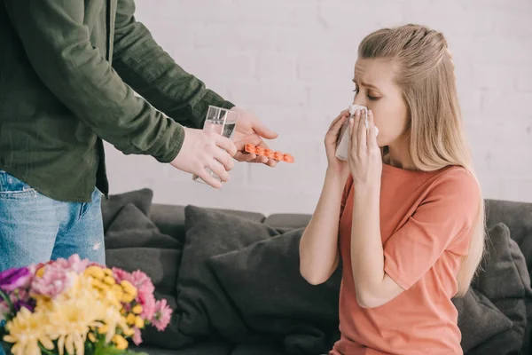 Vista recortada del hombre sosteniendo un vaso de agua y pastillas cerca de la mujer rubia con alergia al polen estornudos en el tejido cerca de las flores - foto de stock