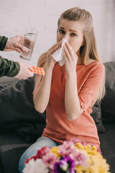 Vista recortada del hombre sosteniendo un vaso de agua y pastillas cerca de la mujer con alergia al polen estornudos en el tejido cerca de las flores - foto de stock