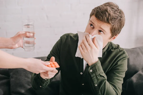 Vista ritagliata della donna che tiene il vetro con acqua e pillole vicino all'uomo starnuti nel tessuto — Foto stock