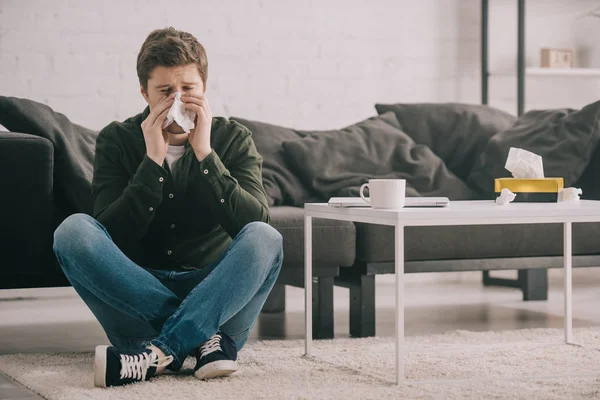 Мужчина сидит на ковре со скрещенными ногами и чихает в тканях возле кофейного столика с чашкой — стоковое фото